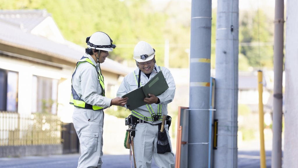 屋外でバインダーの資料を指差し、打ち合わせをする2名の作業員