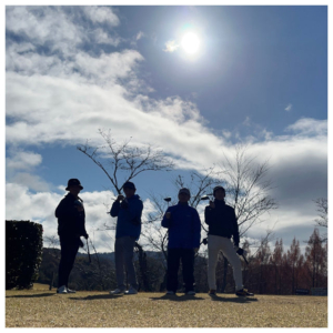 晴れた空の下、ゴルフ場のコース上でクラブを持って仲間たちと並んで立っている田口さん