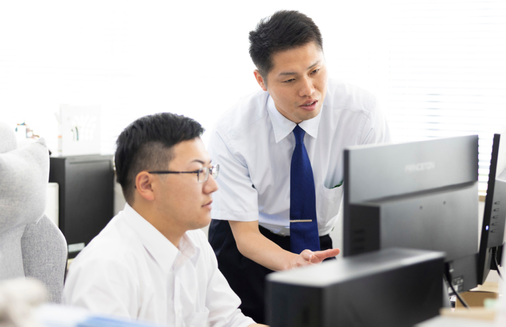 オフィスでパソコンのモニターを指差しながら、同僚と会話をしている田口さん