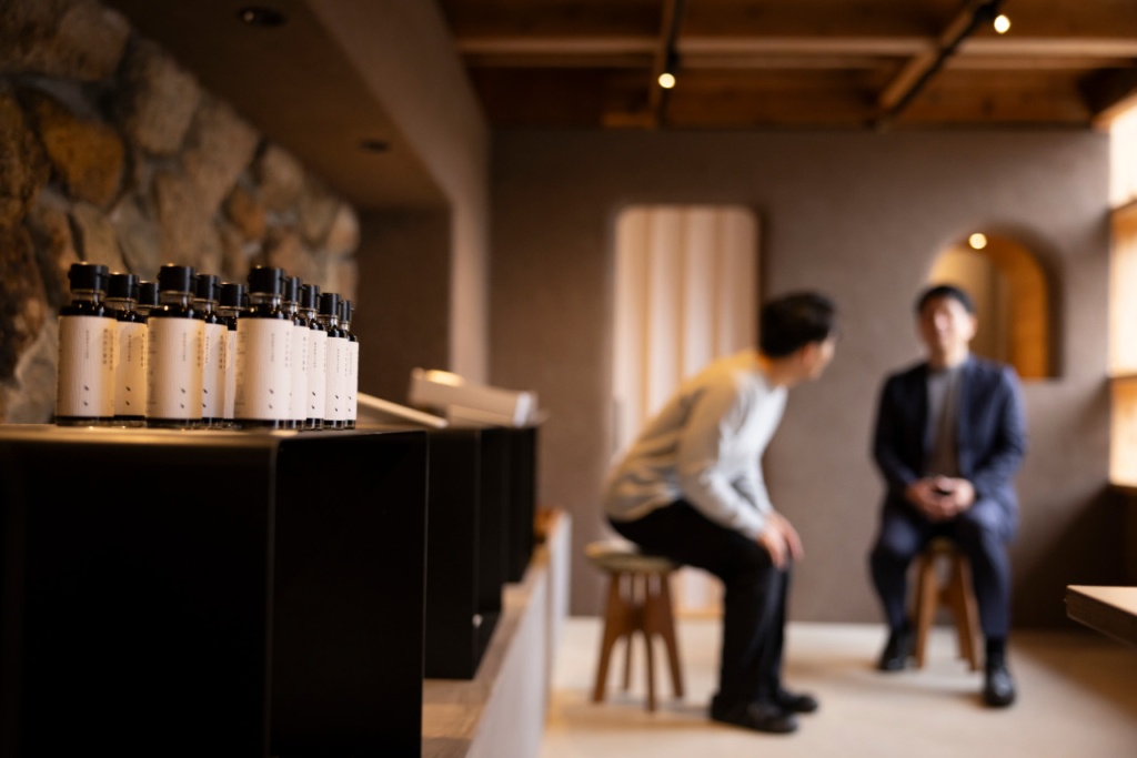 手前に並んだ瓶詰め商品と奥で焦点がぼけて映る羽田さんとインタビュアー