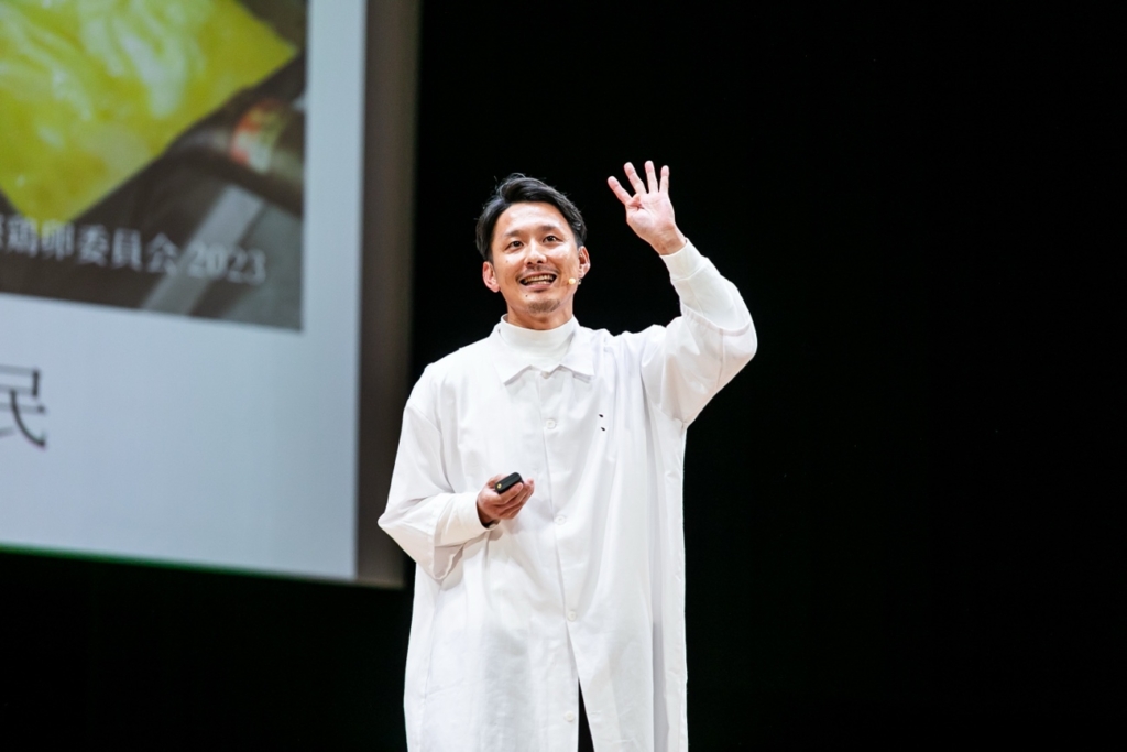ステージ上でマイクを持ちプレゼンテーションを行っている白衣姿の羽田さん