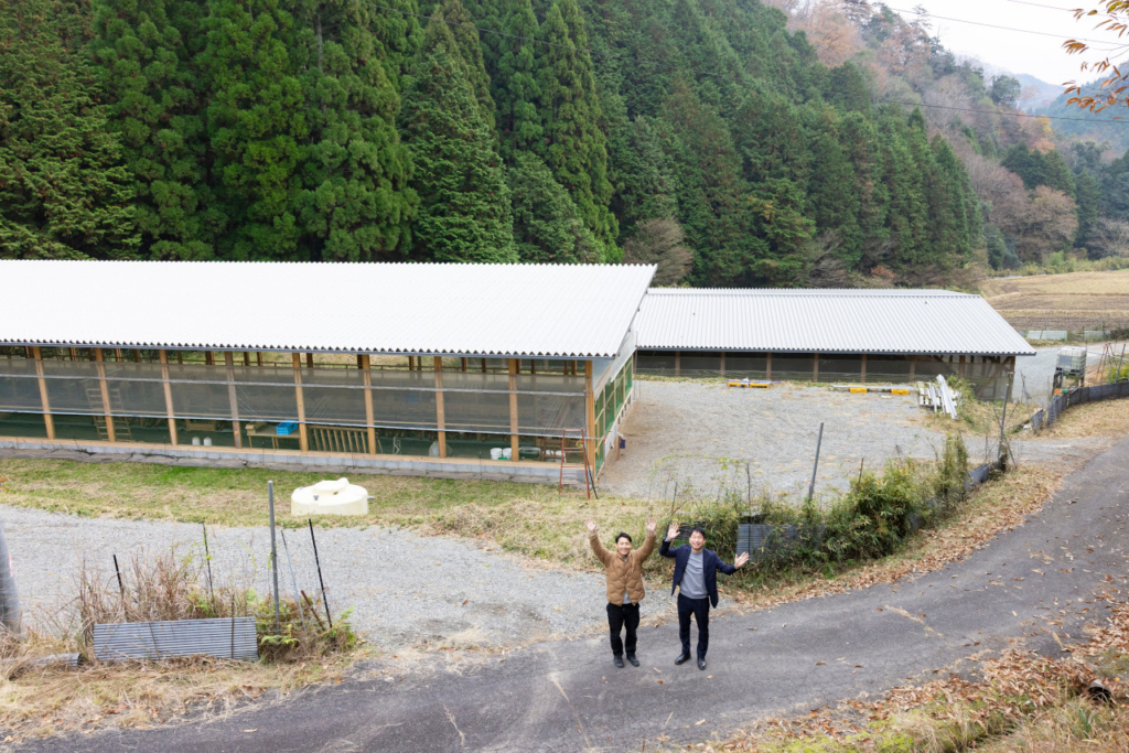 屋外の鶏舎の前でカメラに向かって手を振っている羽田さんとインタビュアー