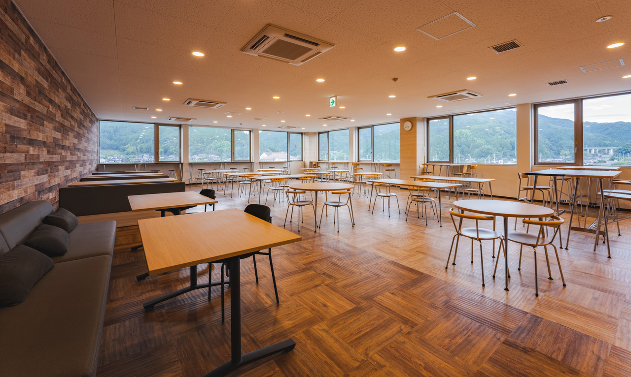 窓から外の景色が見え、複数のテーブルと椅子、ソファが配置された木目調の広い休憩スペース