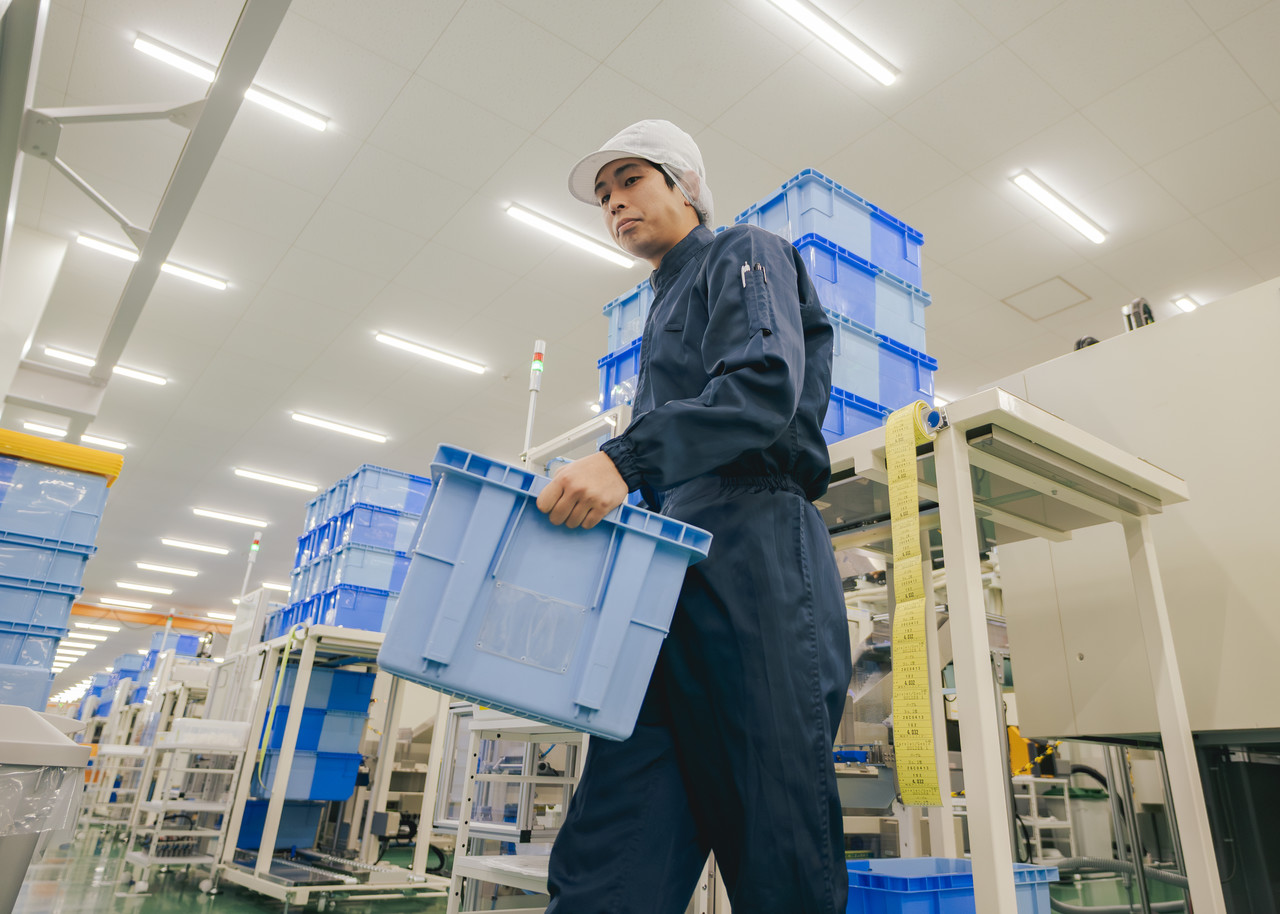 工場内で、製品が入った水色のプラスチックコンテナを両手で持ち運び移動している社員