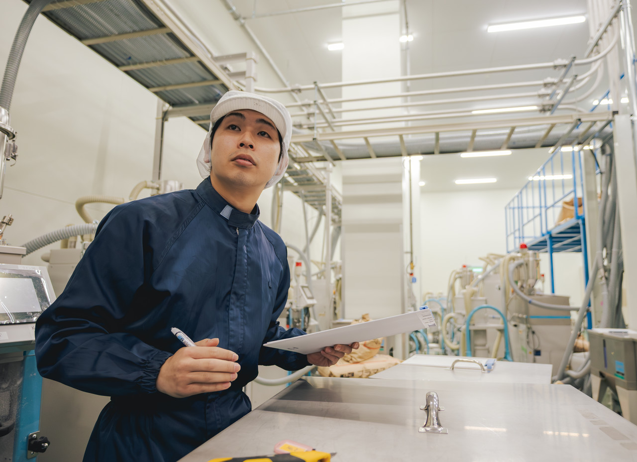 機械の前に立ち、手元のバインダーに挟まれた書類を確認しながら設備の記録をチェックする庄司さん