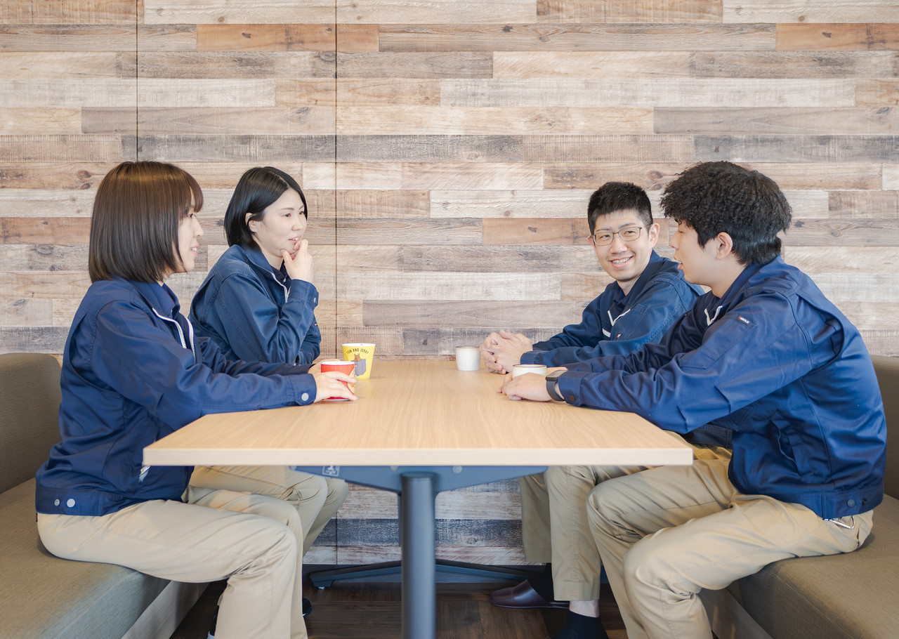 木目調の壁を背景にしたソファ席で、マグカップを片手に4人の社員が向かい合って談笑する様子