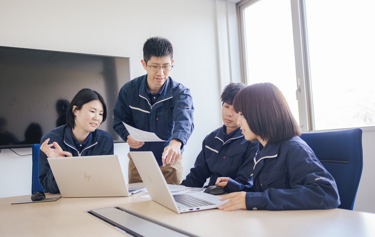 会議室のテーブルで、ノートパソコンの画面を見ながら話し合う作業着姿の4人の社員