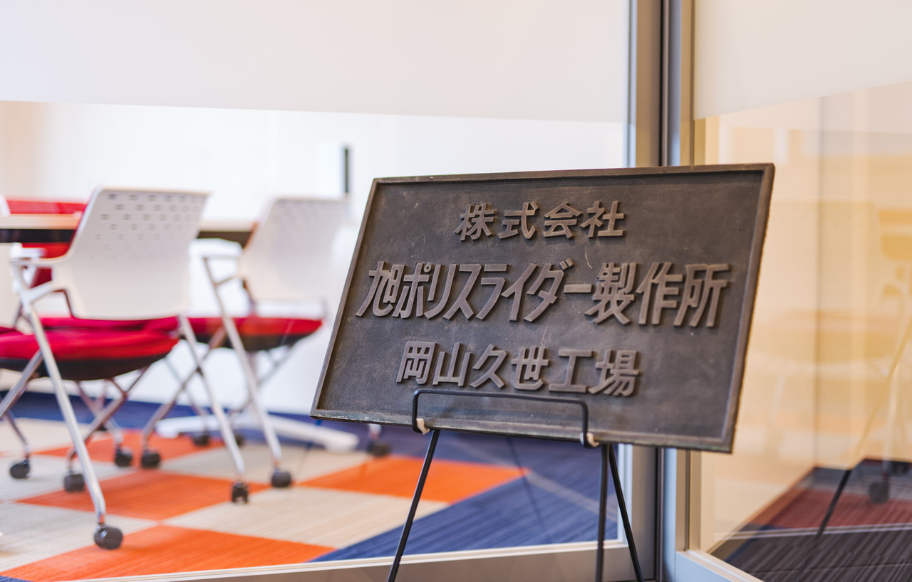 ガラスの仕切りの前に置かれた「株式会社旭ポリスライダー製作所 岡山久世工場」の看板