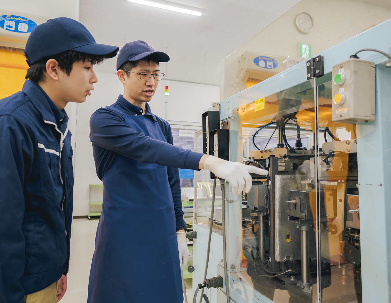 工場内で、先輩社員が成形機を指差しながら、横に立つ後輩社員へ説明をしている様子