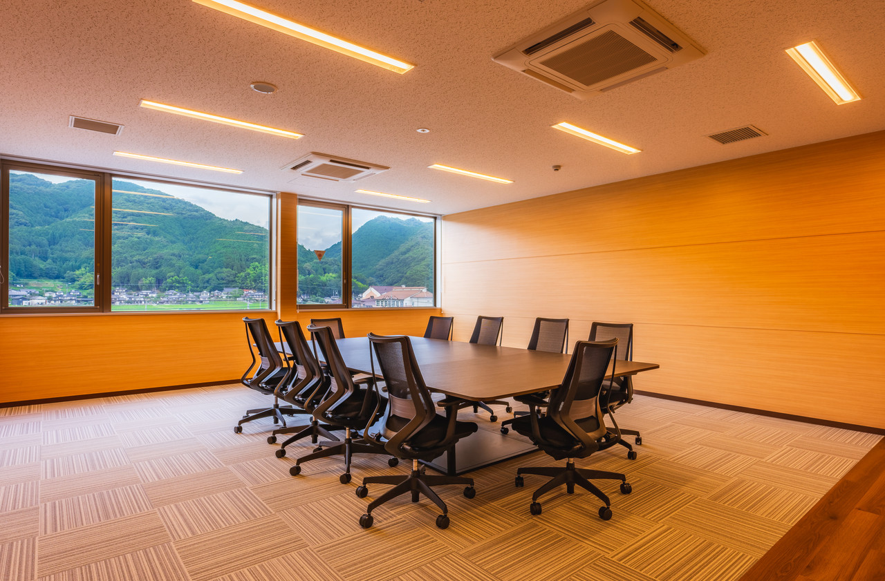 窓から山の景色が見える、木目調の壁と大きなテーブルに椅子が並べられた会議室
