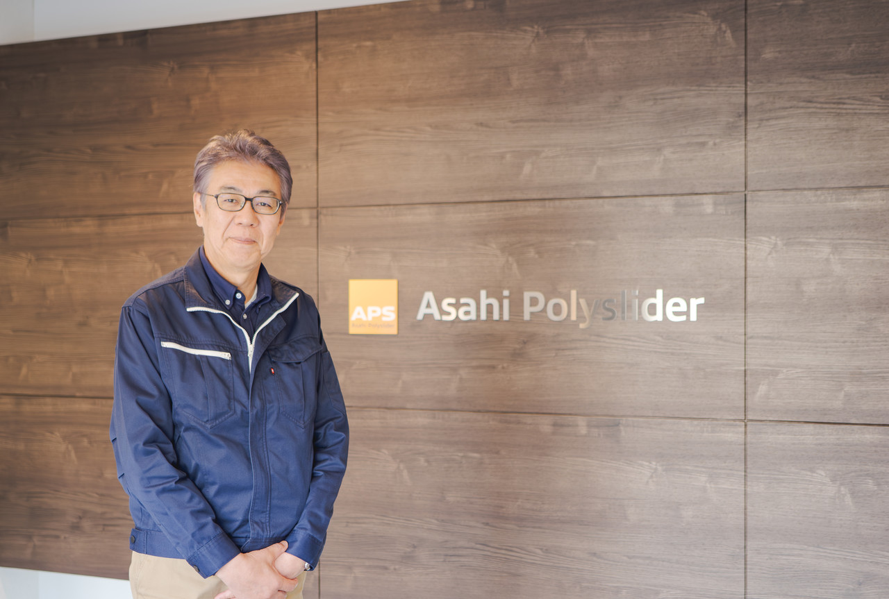 企業ロゴが掲示された木目調の壁の前に立つ、作業着姿の太田哲也代表取締役社長