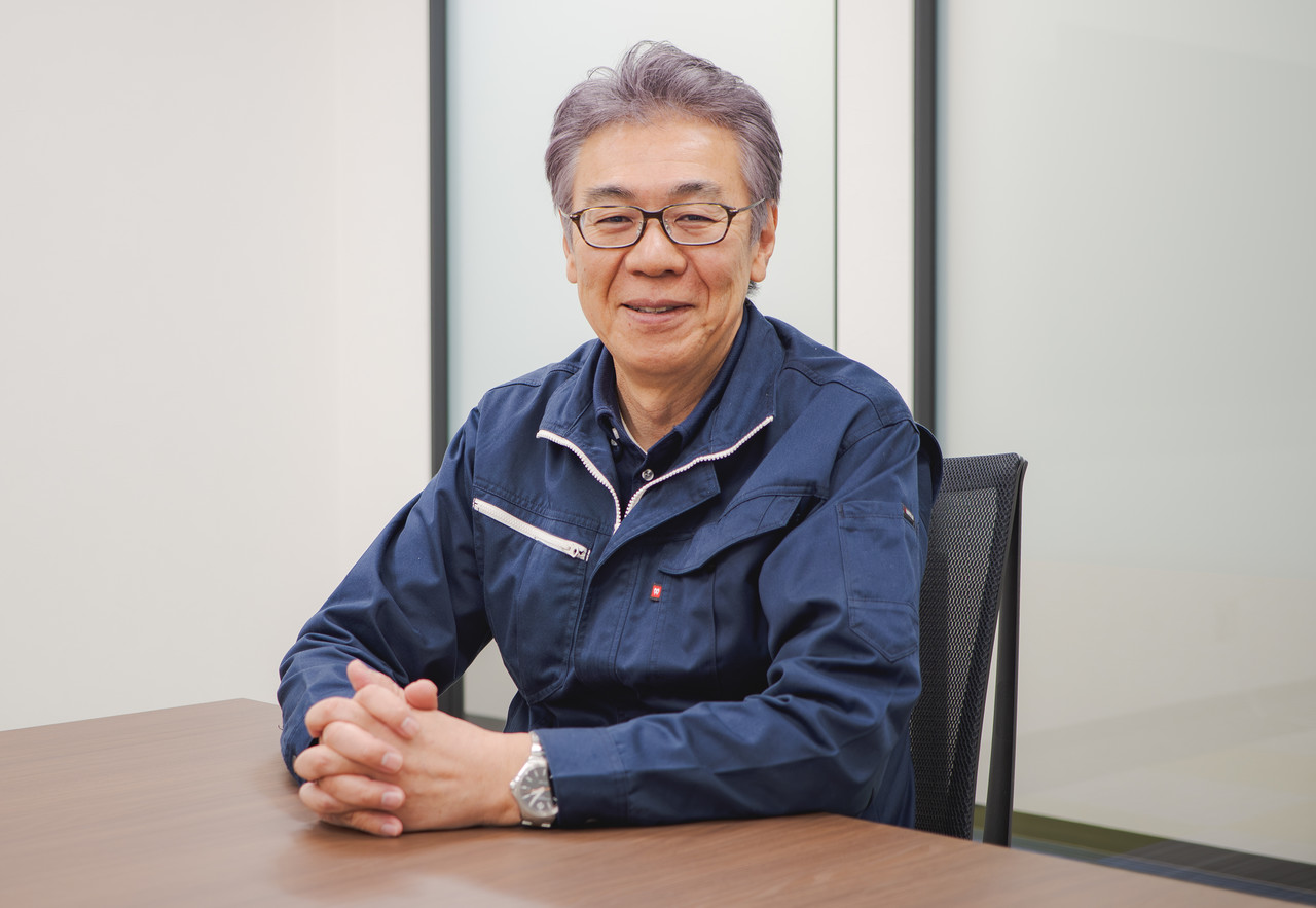 会議室のテーブルに座り、両手を組んでカメラに視線を向ける作業着姿の太田哲也社長