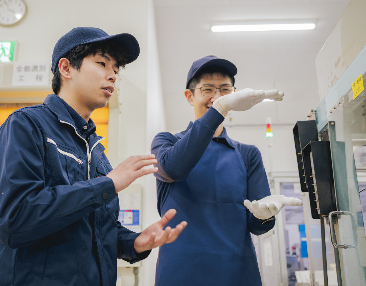 工場内で、機械を前にして身振りを交えながら先輩社員とコミュニケーションを取っている庄司さん