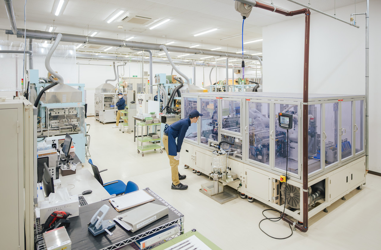 複数の機械が並ぶクリーンな工場内で、透明なカバーで覆われたライン設備を確認する社員