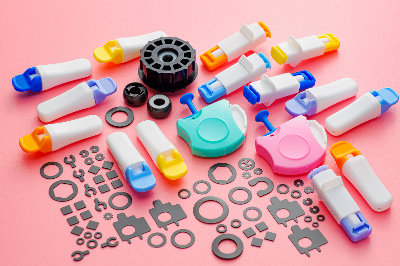 ピンク色の背景に並べられた、医療用採血器具や黒い精密部品など様々なプラスチック製品