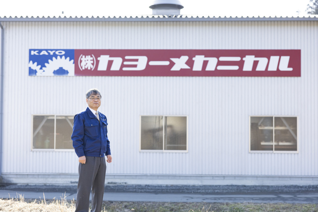 カヨーメカニカルのロゴが壁面に大きく掲げられた工場の前に立つ小虎修一さん