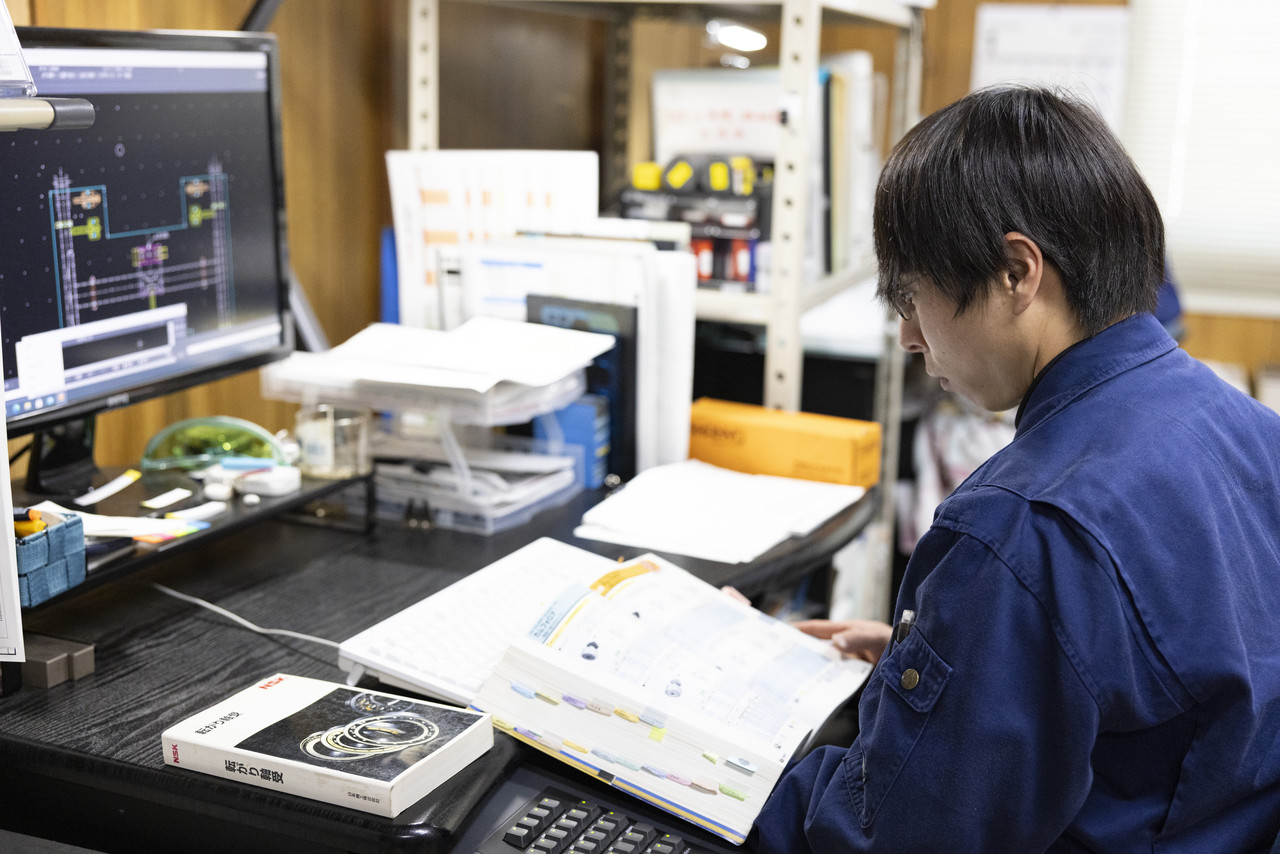 デスクで、パソコンの画面を開きながら分厚い書籍を見ている上鶴さん