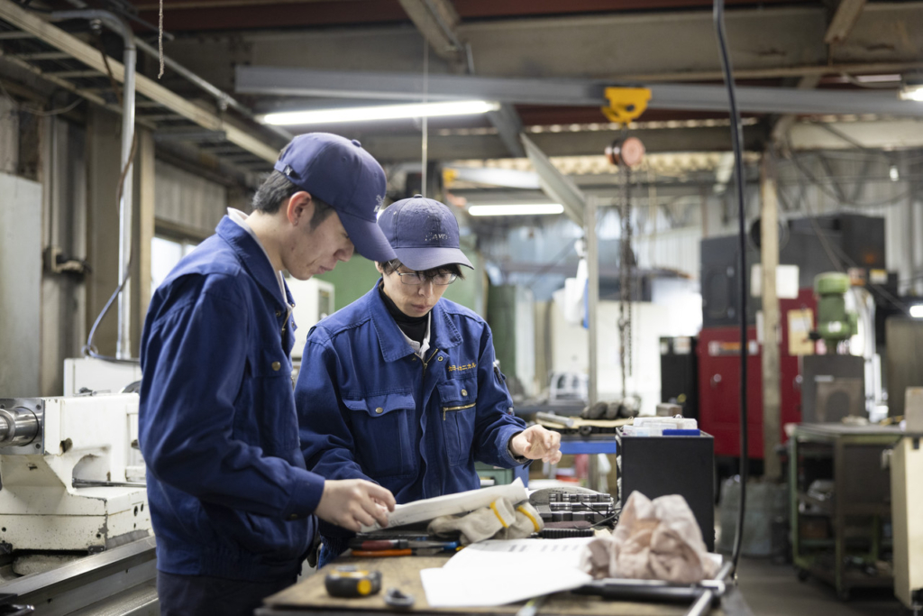 図面を確認しながら打ち合わせをする青い作業服の男性社員2名