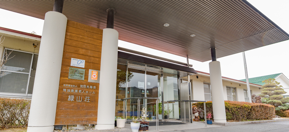 大きな庇と白い円柱、施設名が刻まれた木の看板がある緑山荘の正面玄関。