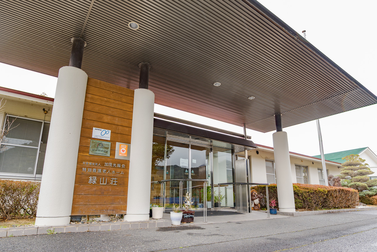 大きな庇と白い円柱、施設名が刻まれた木の看板がある緑山荘の正面玄関。