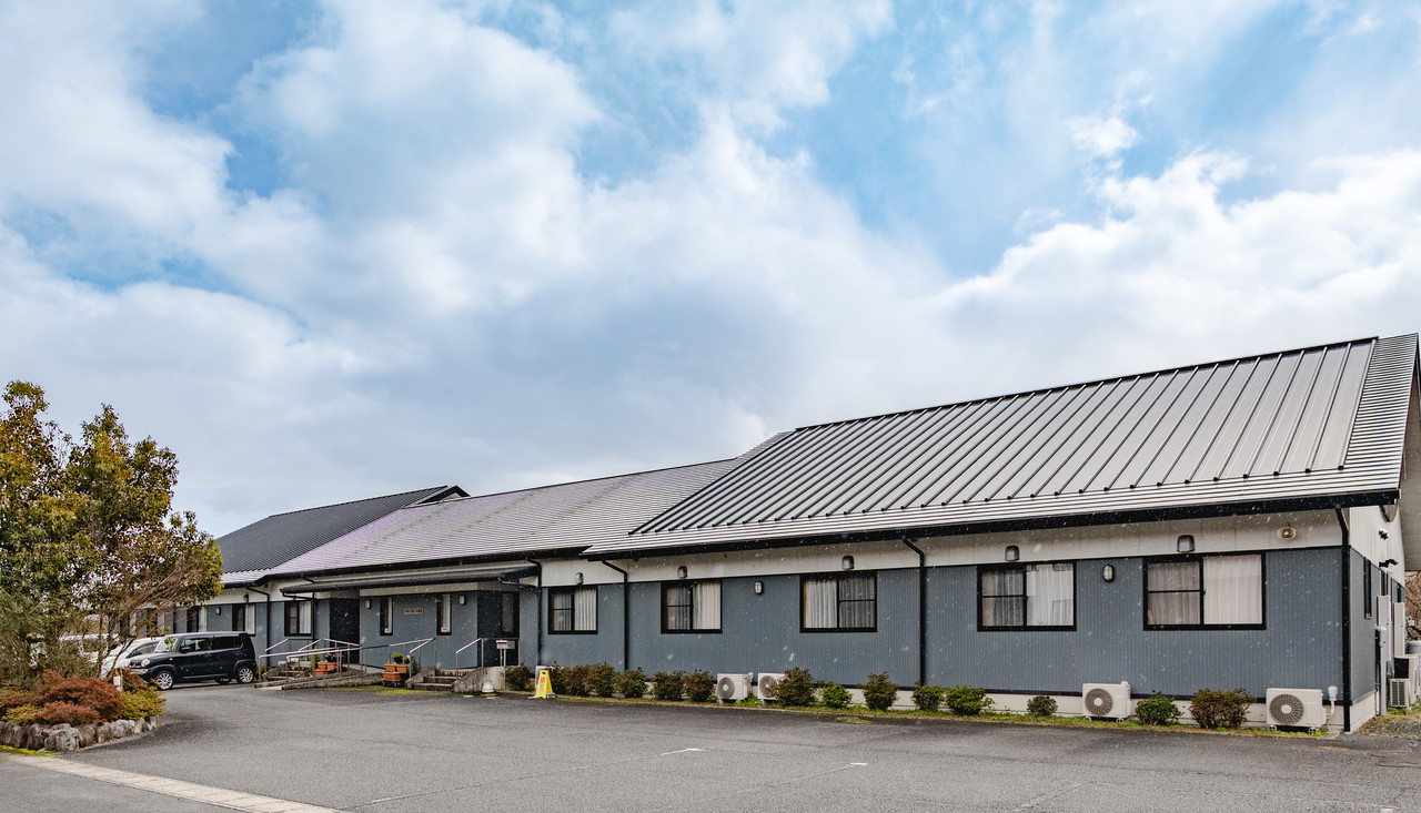 青い外壁と大きな勾配屋根が特徴的な、平屋建てのグループホーム杉宮の外観と駐車場。