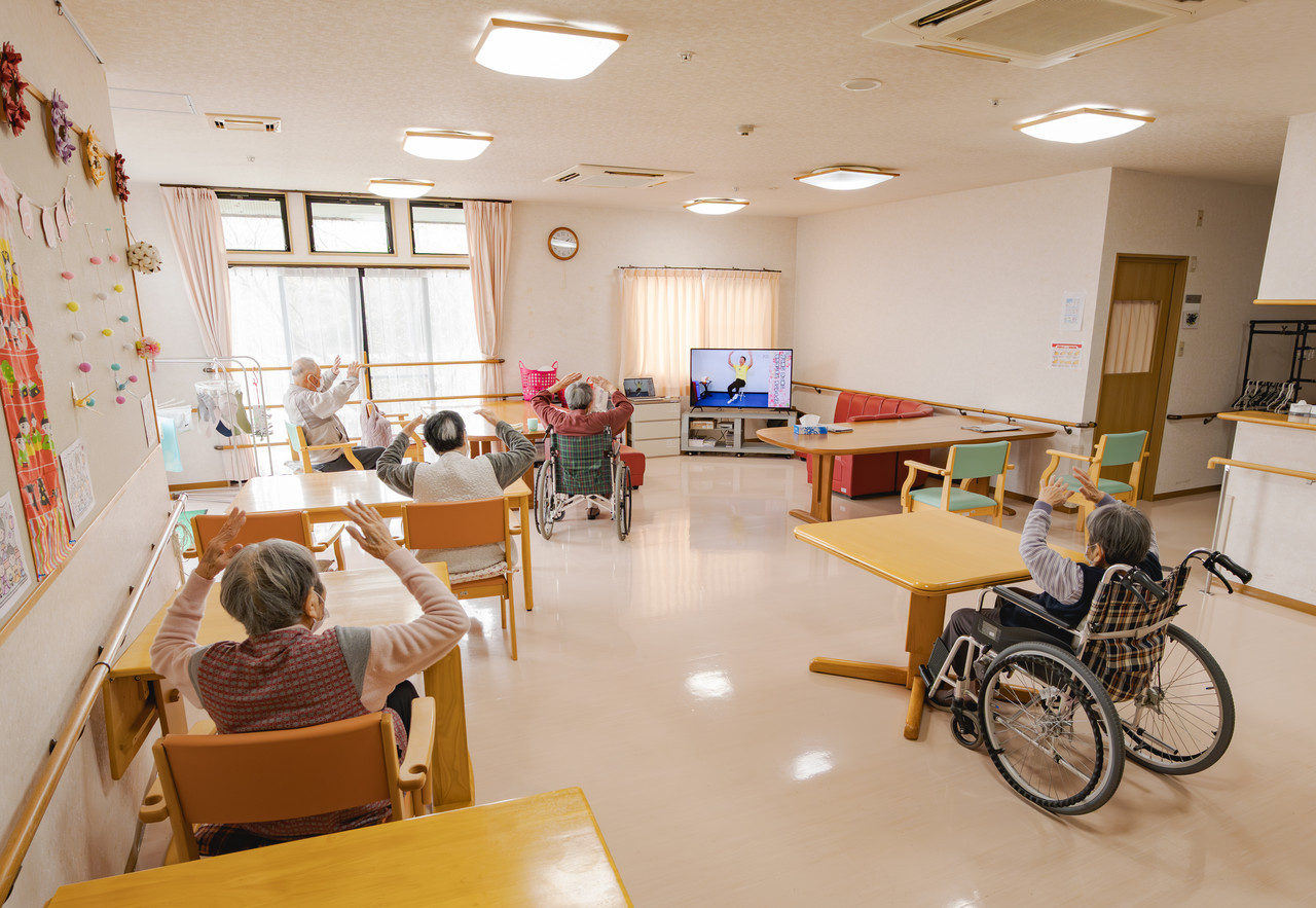 ホールの大型テレビに映る映像に合わせて、椅子に座りながら腕を動かす体操をする利用者。