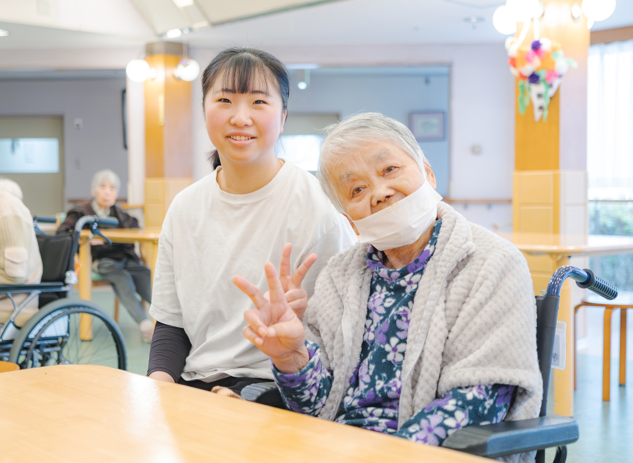 井上さんが車椅子に座った高齢の利用者と並んで、2人でピースをしてこちらを見ている様子