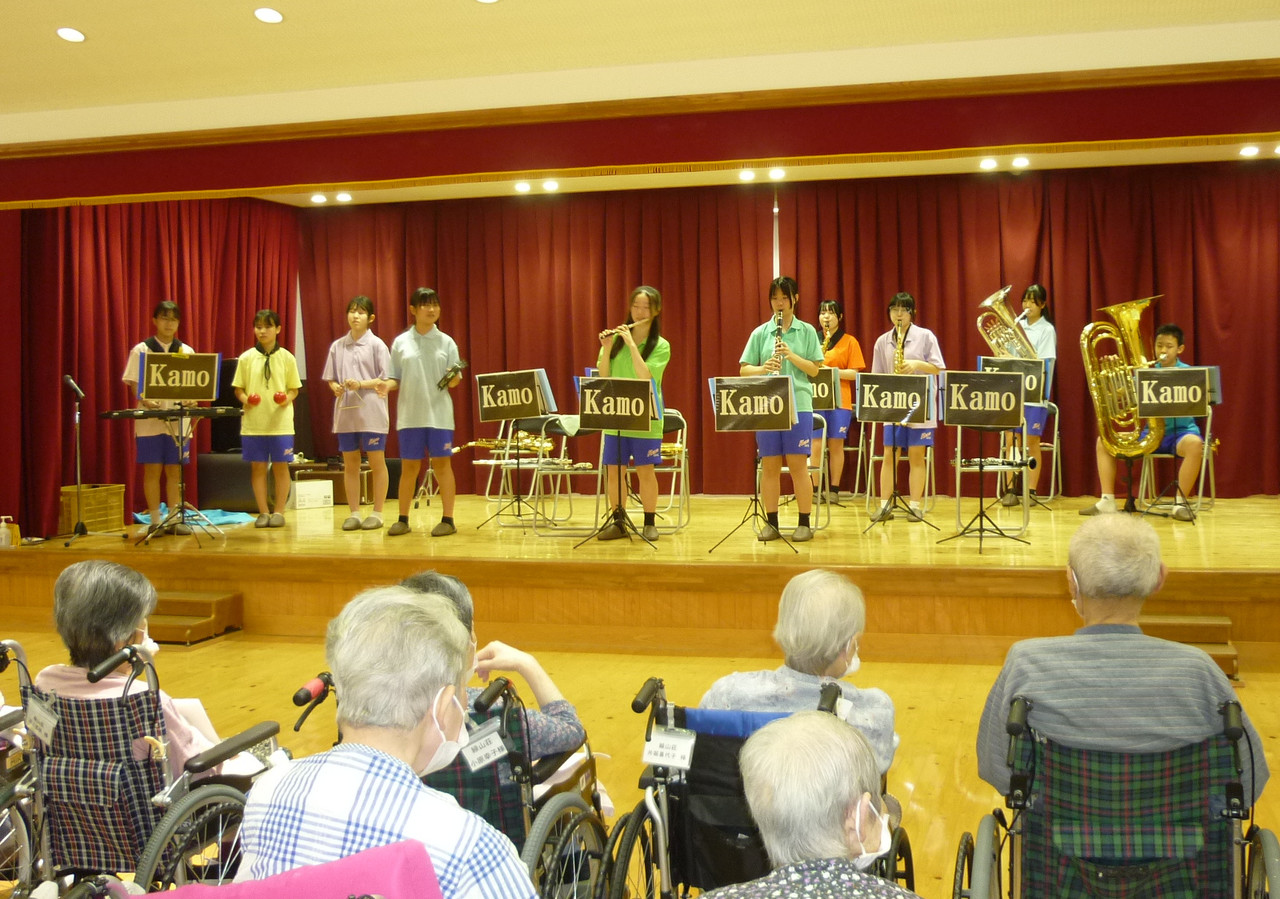 施設のステージで、吹奏楽の楽器を演奏する地元中学校の生徒たちと、鑑賞する利用者たち。