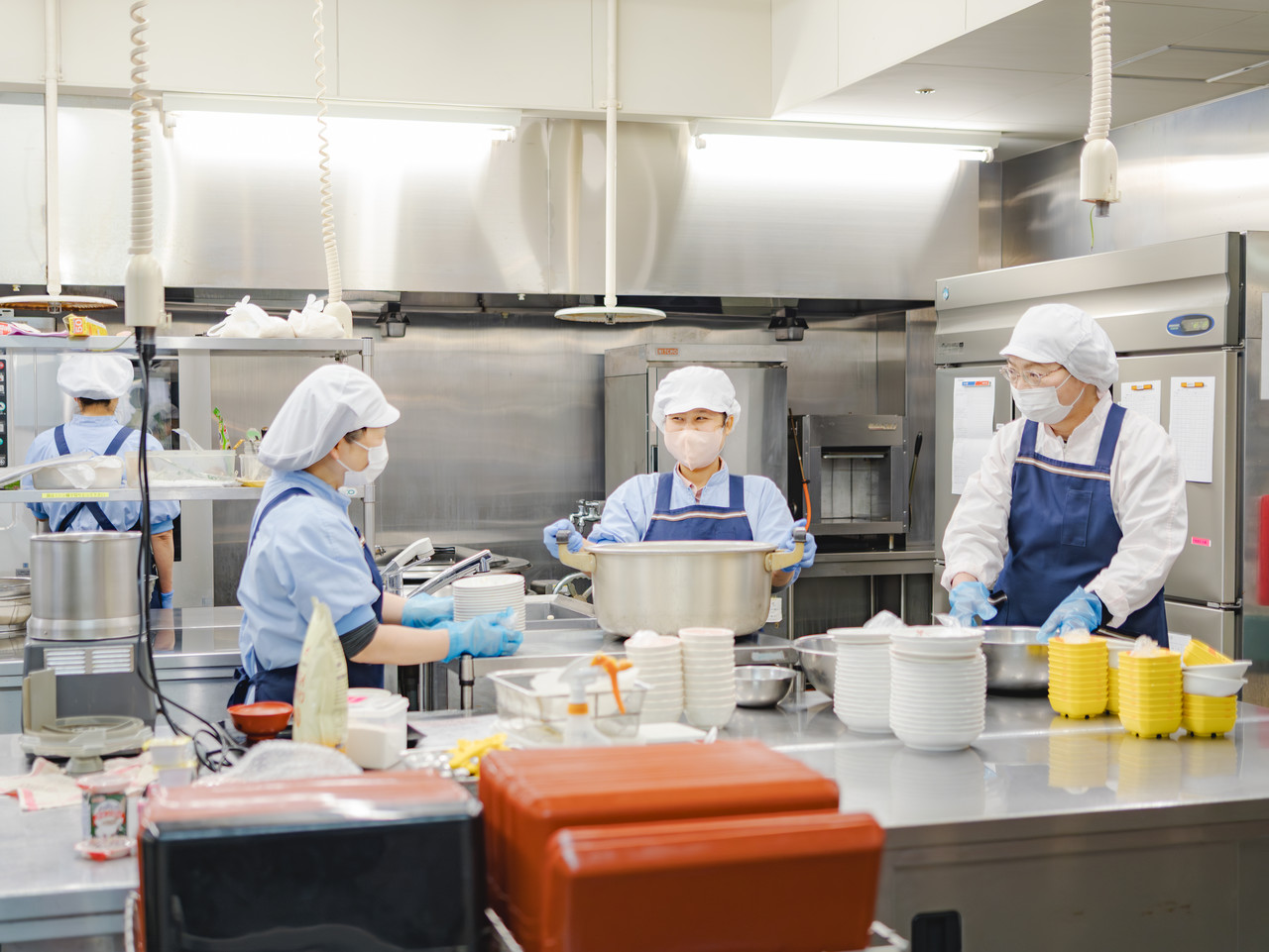衛生服とマスクを着用したスタッフが、大きな鍋や食器を使用して食事の準備をする調理室。