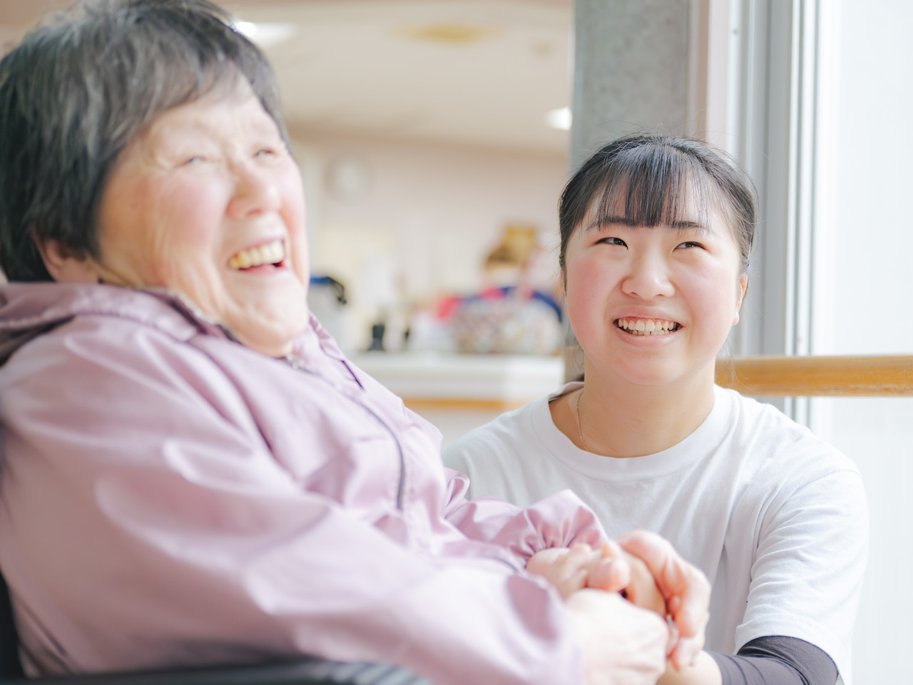 スタッフに手を握られ、楽しそうに笑い合う車椅子の高齢女性利用者と笑顔のスタッフ。