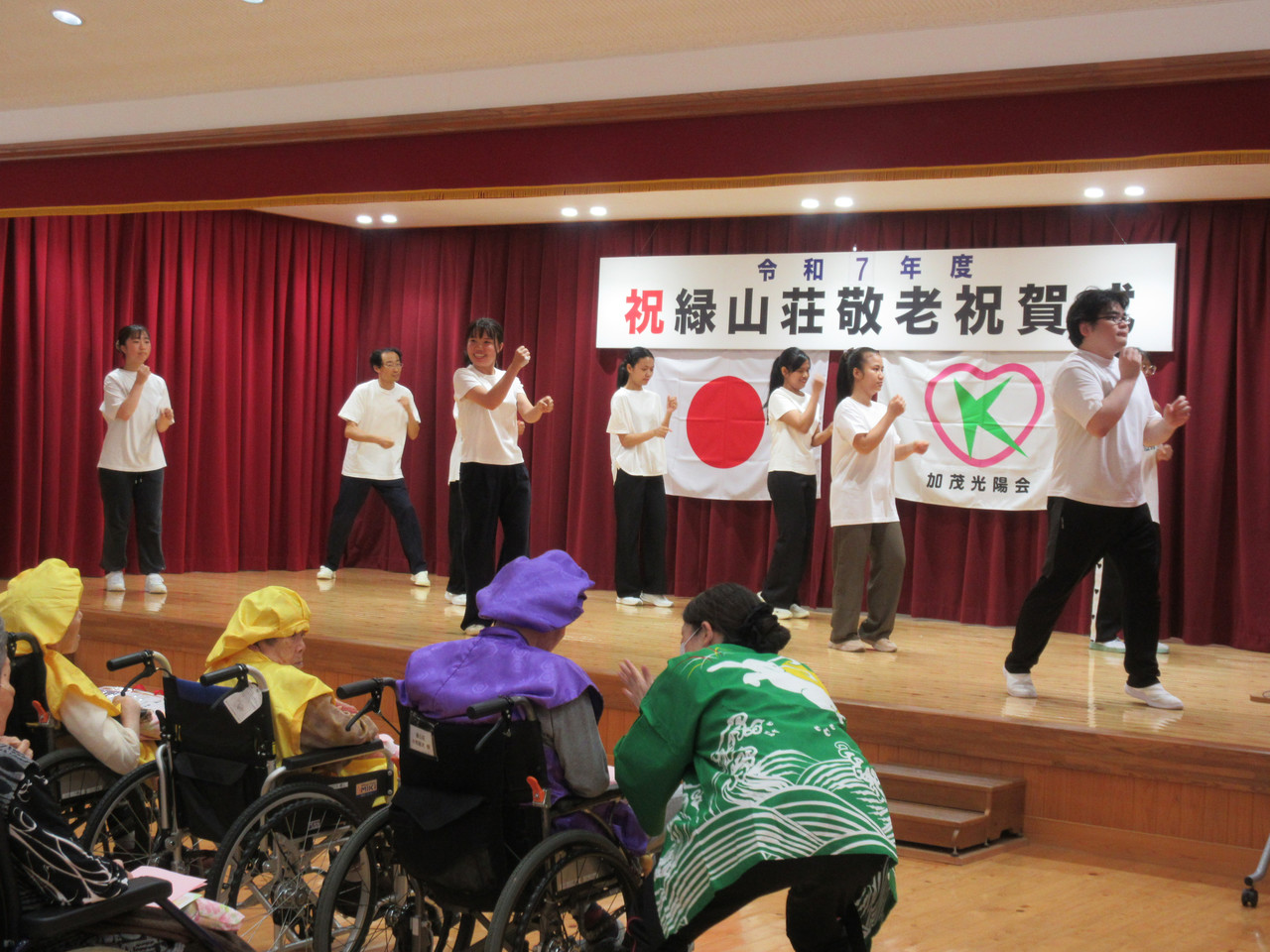 敬老祝賀会のステージ上で、白いTシャツと黒いパンツを着用してダンスを披露するスタッフ。