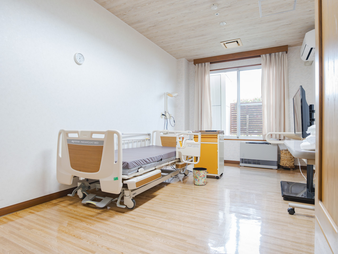 ベッド、テレビ、収納家具が完備された、木目調の床と白い壁の明るい一人用個室。