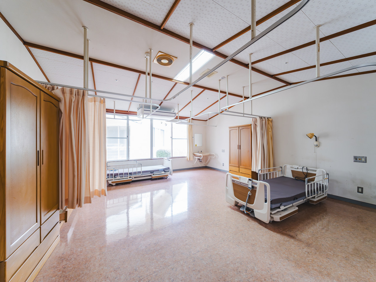 窓際にベッドが配置され、木製の収納家具とカーテンの仕切りがある清潔なグループ居室。