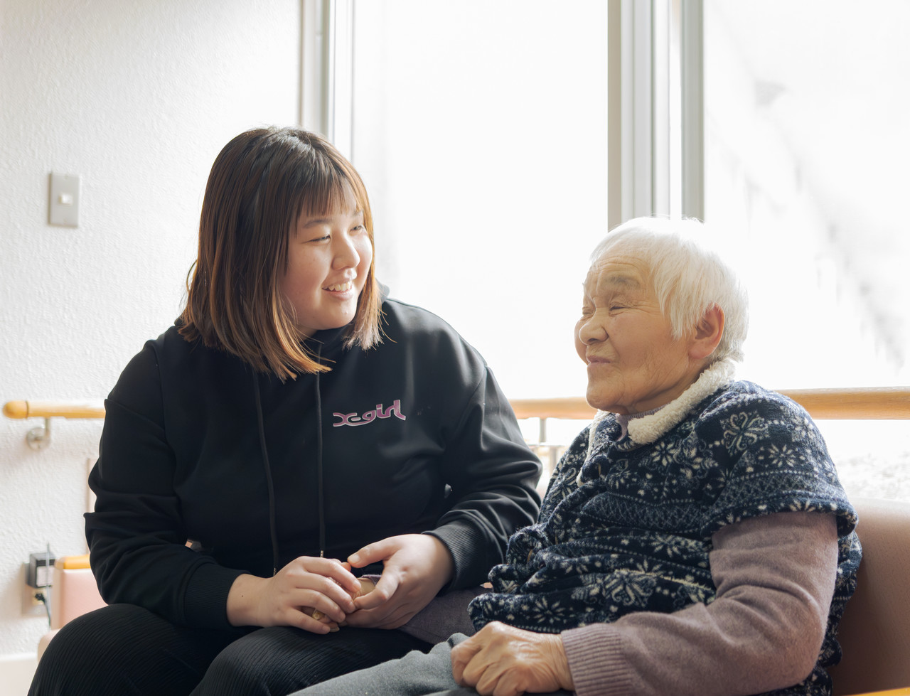 介護スタッフが高齢の利用者と手を握り合い、お互いに笑顔で会話している様子