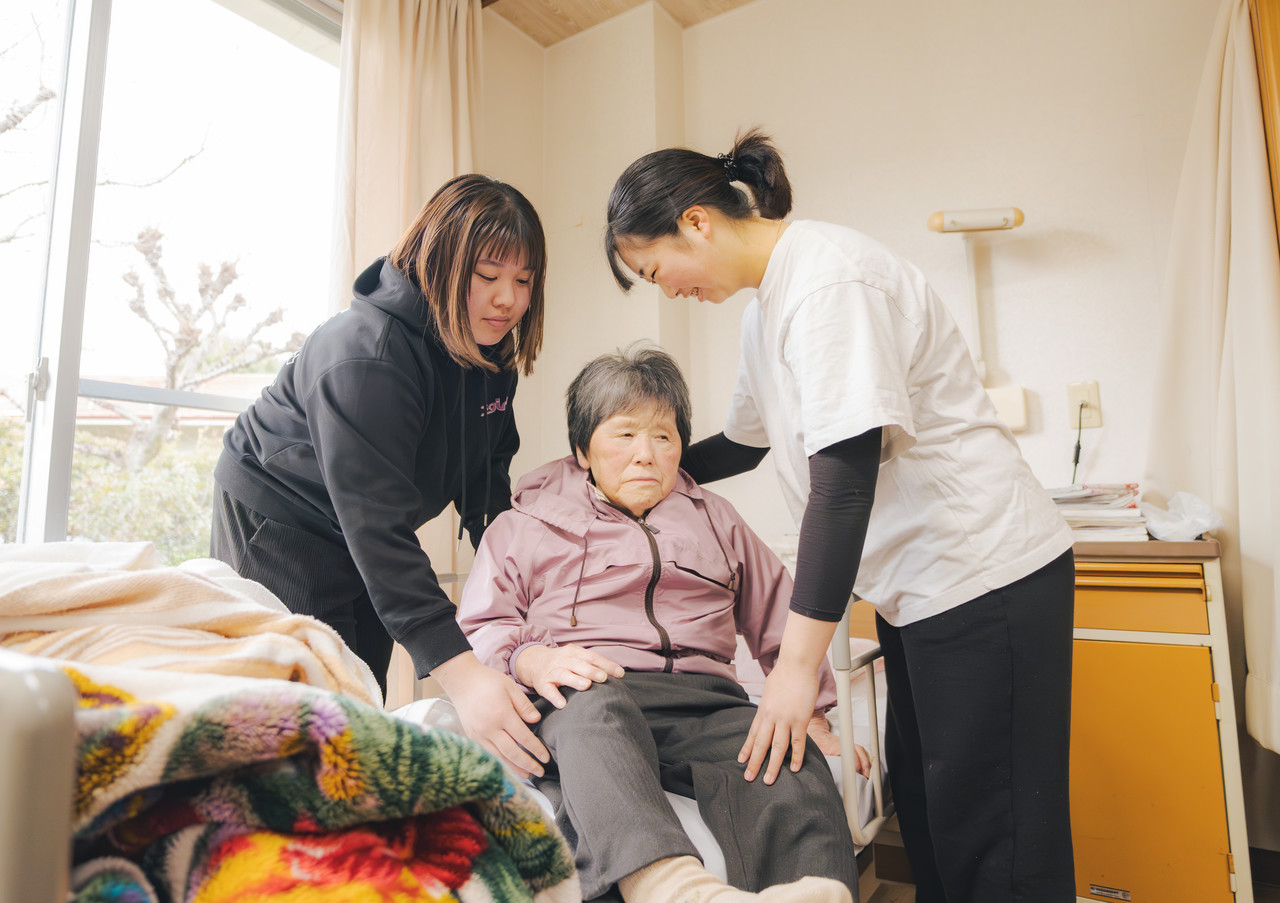 複数の介護スタッフが協力して、ベッドにいる利用者の離床介助を安全に行っている様子