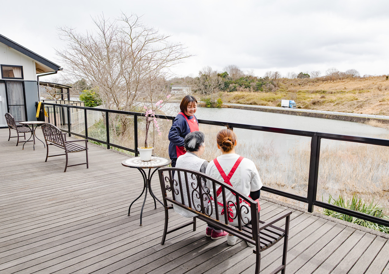 テラスのベンチに座る利用者と芳本さんに、職員が話しかけている様子