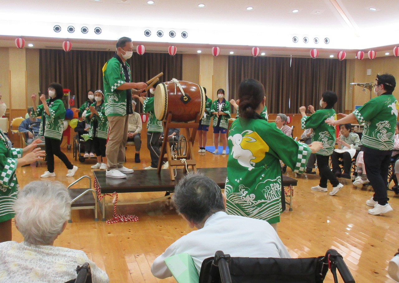 納涼祭の会場で、緑の法被を着たスタッフが和太鼓を叩き、その周りで踊りを披露する様子。