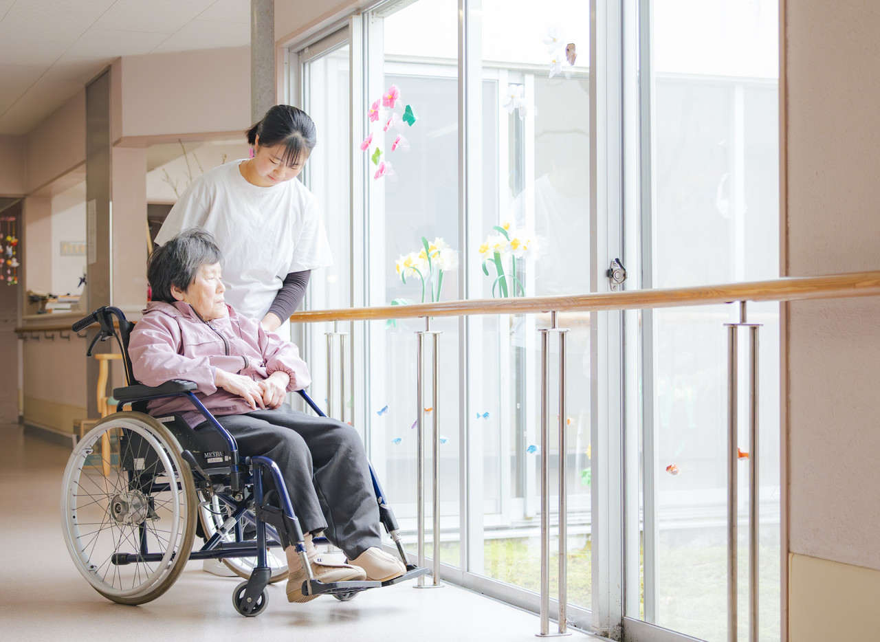窓から光が差し込む廊下で、車椅子の利用者に寄り添いながら歩行の介助を行うスタッフ。
