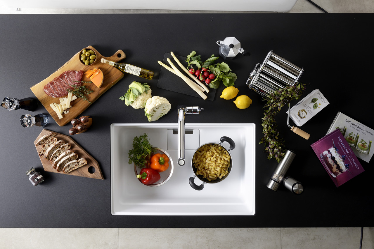 黒いキッチンカウンターの上で、パスタ、肉、野菜などの食材や調理器具が配置されている様子を真上から撮影した写真。