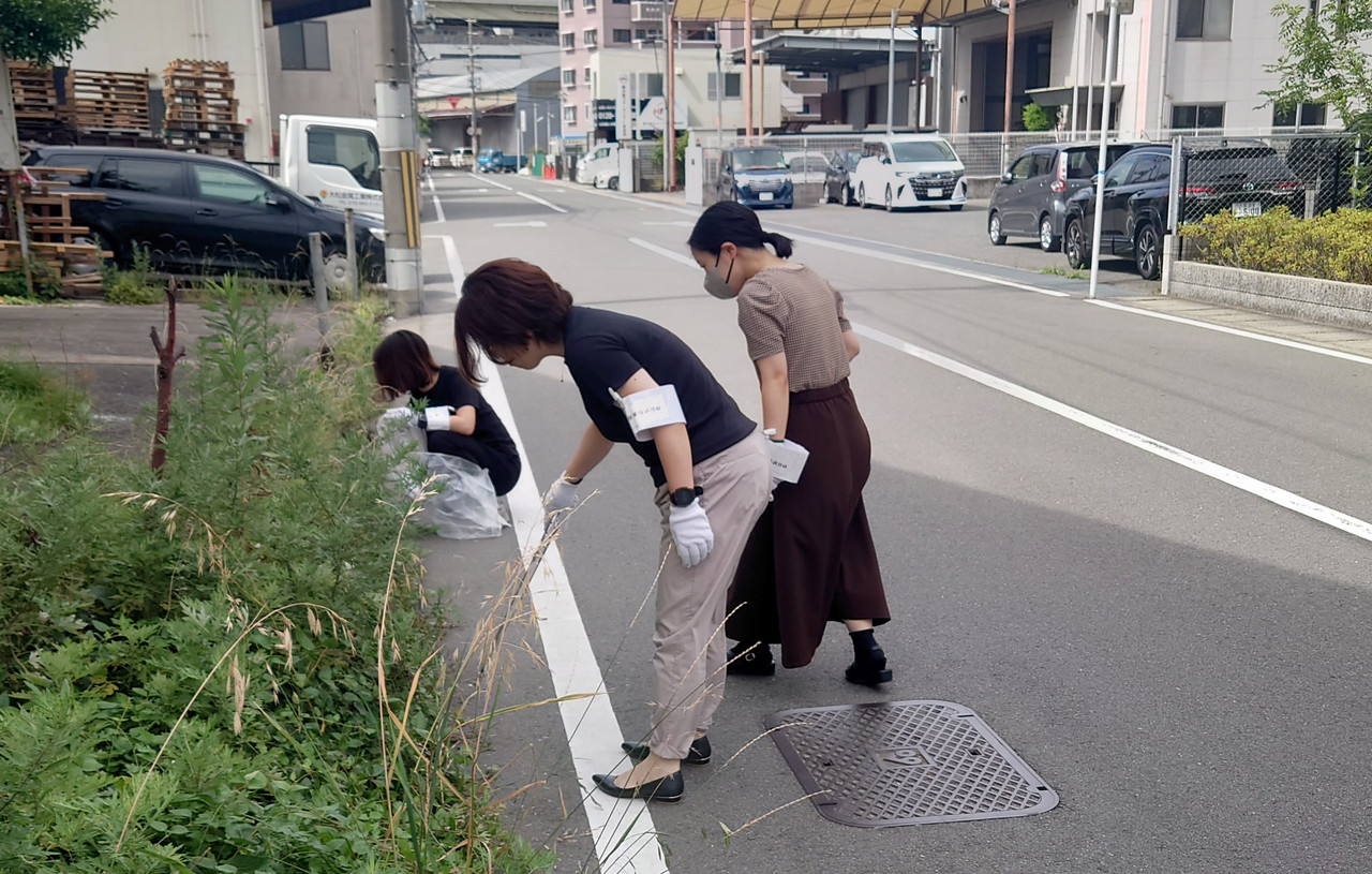工場周辺の道路脇で、従業員が軍手をはめて草むしりやゴミ拾いなどの清掃活動を行っている様子。