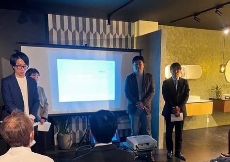 ショールーム内でプロジェクターのスクリーンを背景に、スーツやジャケット姿の従業員が立って発表を行っている様子。