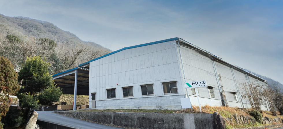 山を背景に建つ、トリムス岡山第3工場の白い建物の外観。工場の前には看板が立っており、その手前にはスロープがある。