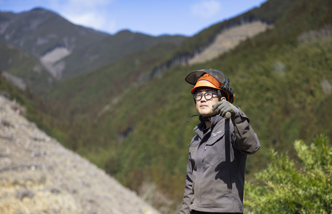 伐採後の山林を背景に、道具を手に持ち遠くを見つめる作業員