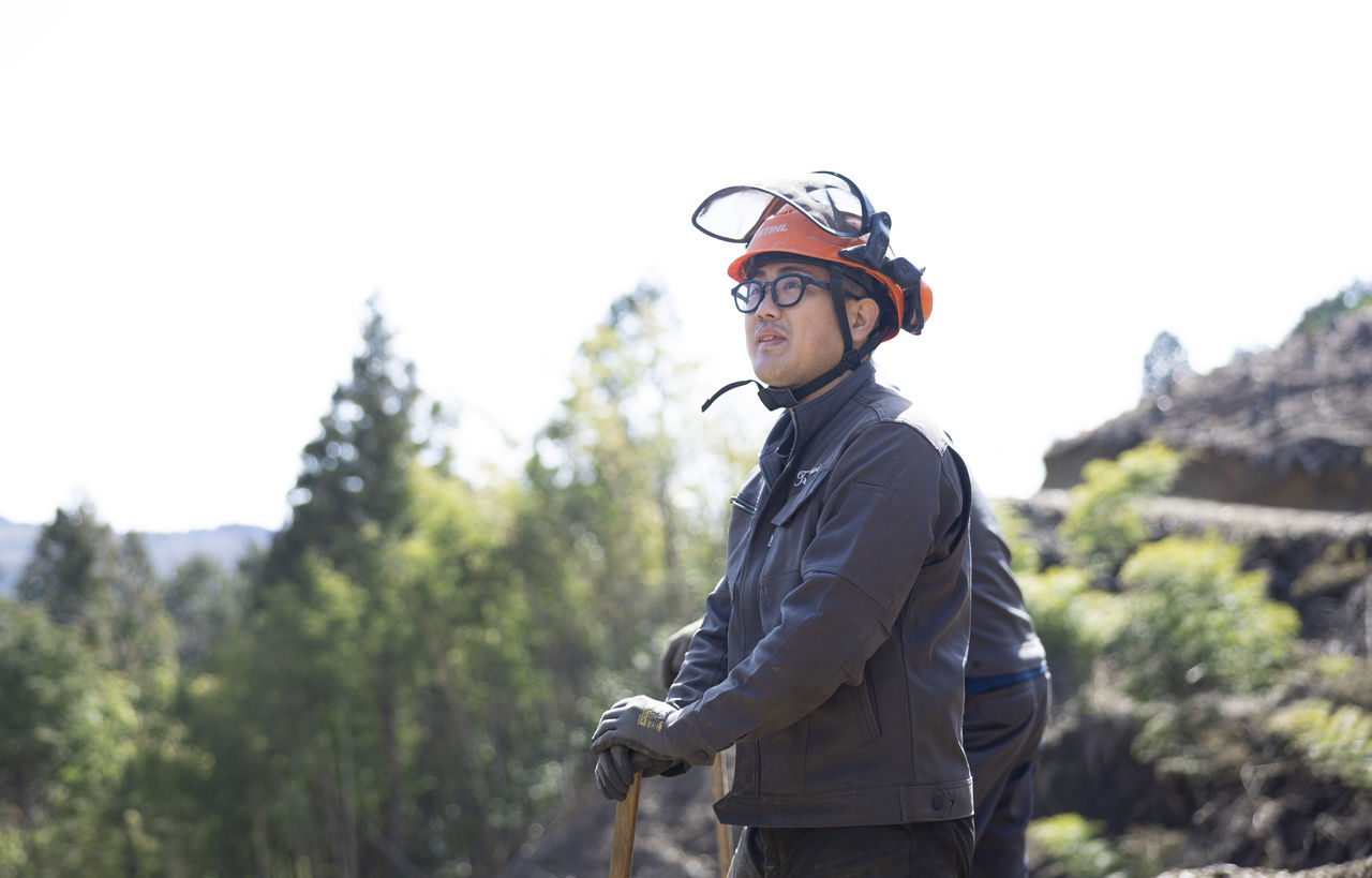 クワを手に持ち、伐採後の山林で遠くを見上げる三宅さん