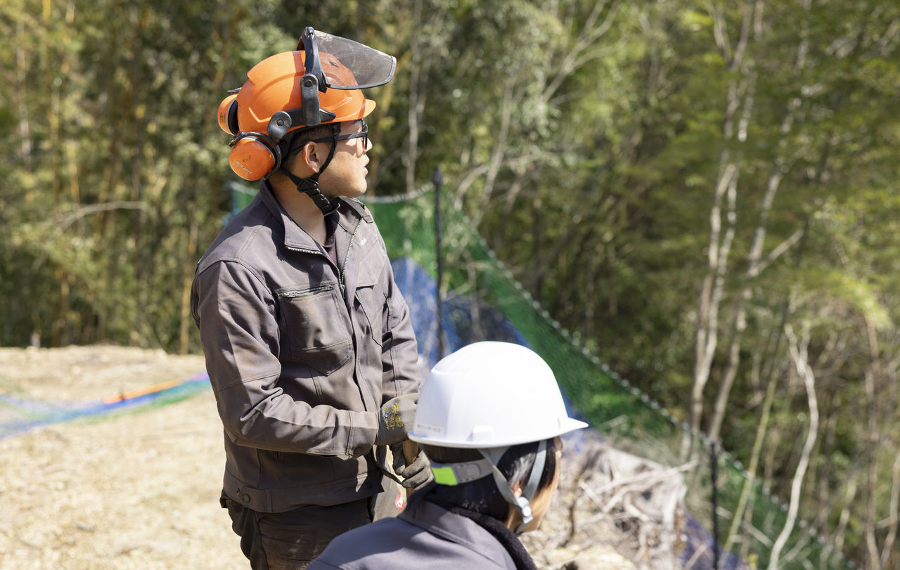 鹿よけネットが張られた斜面を背に、作業の様子を確認する三宅さんともう1人の作業員