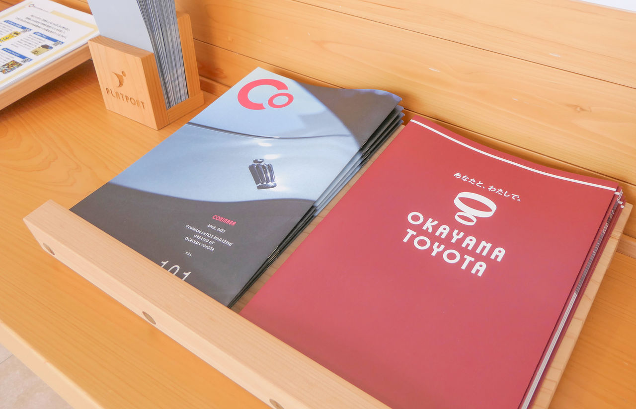 木製のスタンドに置かれた2種類の冊子。左側は車の写真が表紙の「COMIMAGA」、右側はえんじ色に白いロゴの「OKAYAMA TOYOTA」のパンフレット。