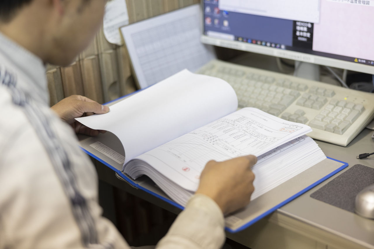デスクに座り、パソコンのモニターの前に置かれたファイルに綴じられた検査書類を確認する従業員。