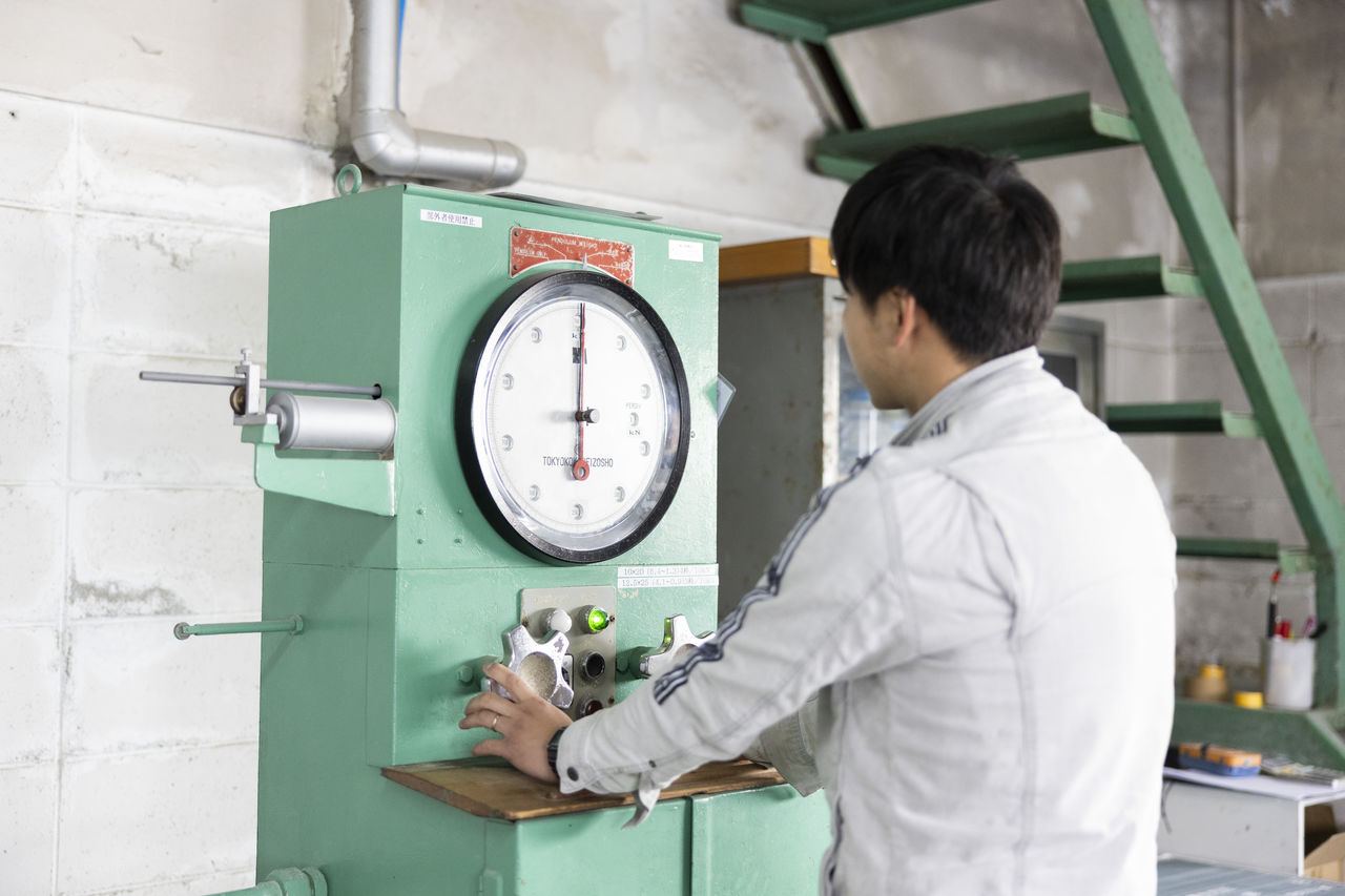 工場内に設置された、目盛りのある大きな緑色の検査機器を操作している従業員。