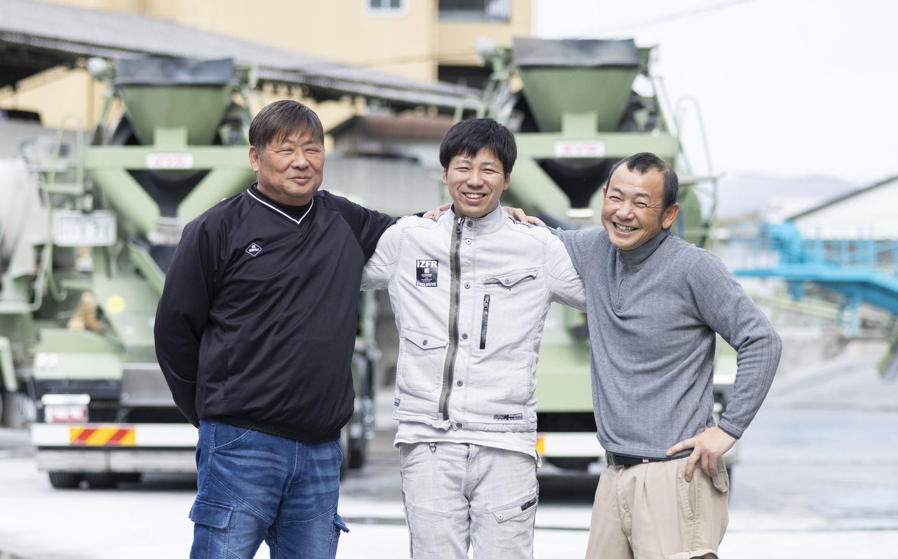 工場の敷地内で、ミキサー車を背景に先輩社員2名と肩を組んで笑顔を見せる大寺さん。