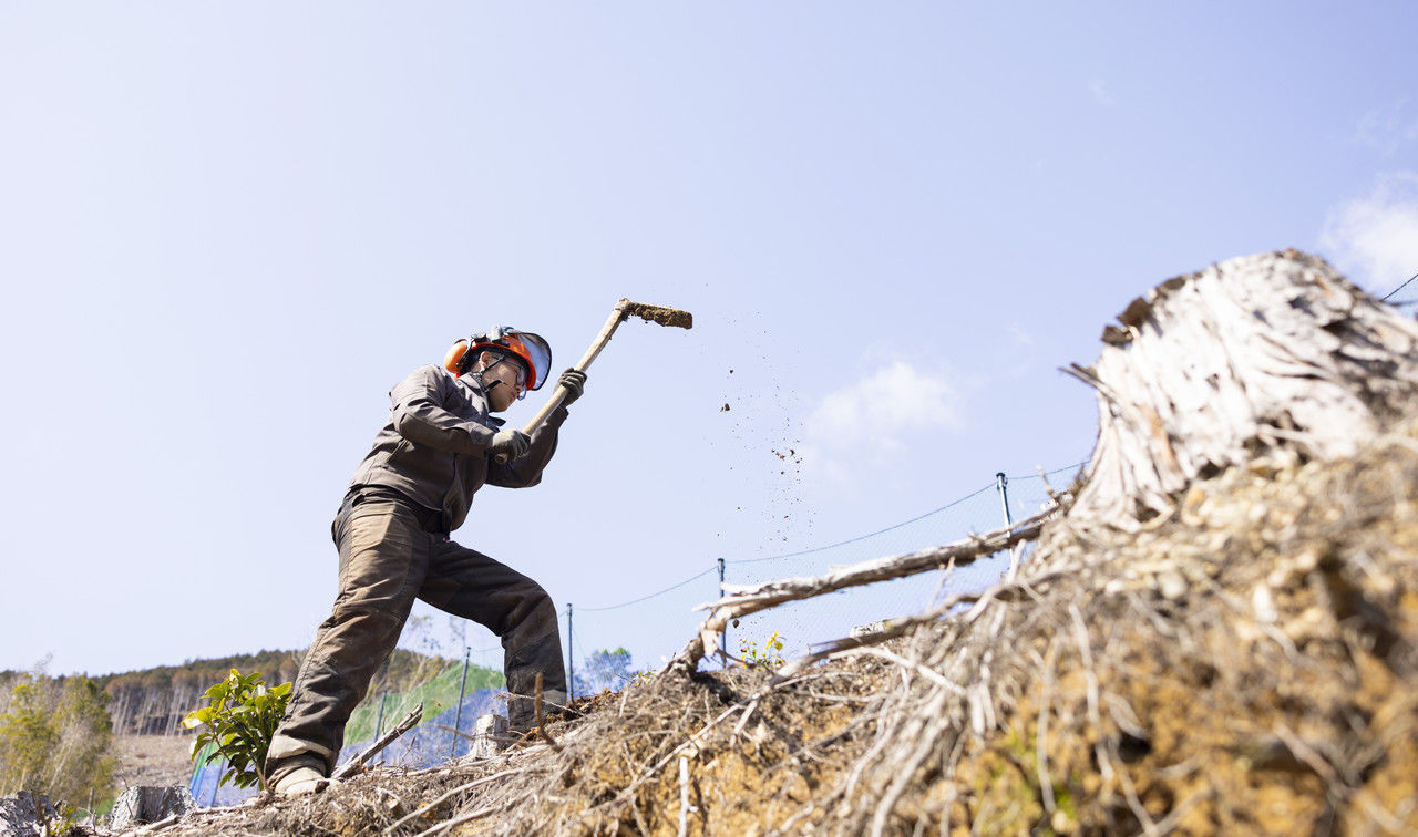 青空の下、山の斜面でクワを振り上げて作業を行う作業員